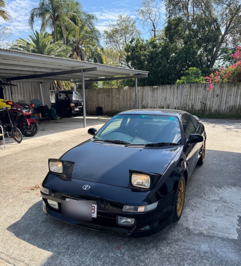 Black 1997 Toyota MR2 with its pop-up headlights raised; number plates blurred.